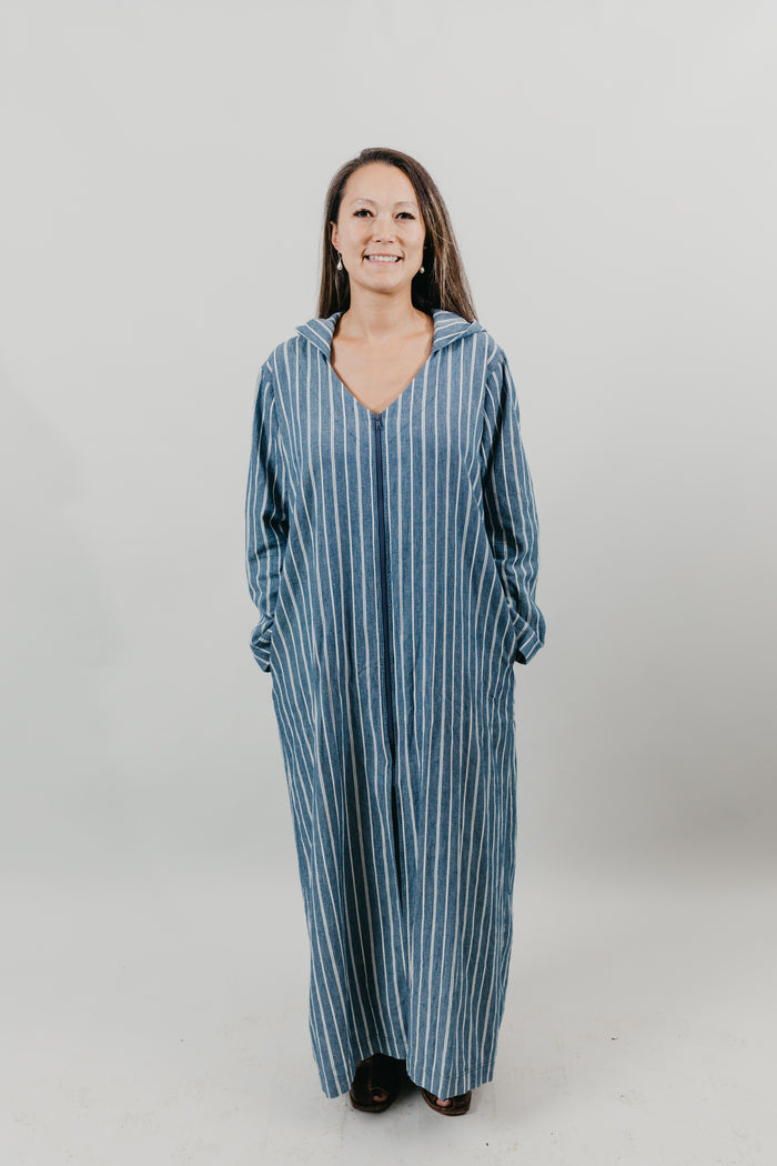 Asian woman wearing a floor length blue and white vertical striped Djellaba with her hands in her pockets. Background is white back drop.