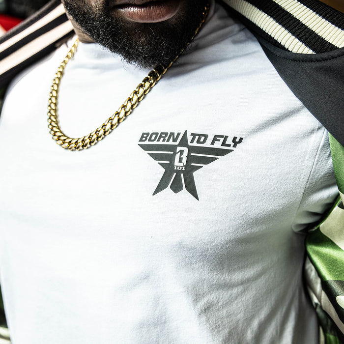 Close up white t-shirt with black “BORN TO FLY” lettering, black star, and white BL101 logo inside the star positioned on the top corner of the shirt on person