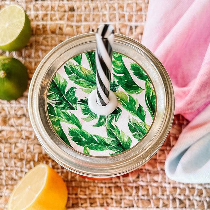 Mason jar straw lid with silver ring and a white sticker that has palm leaves in print. Photographed as a flat lay in a weave mat with a scarf and some lemon slices.