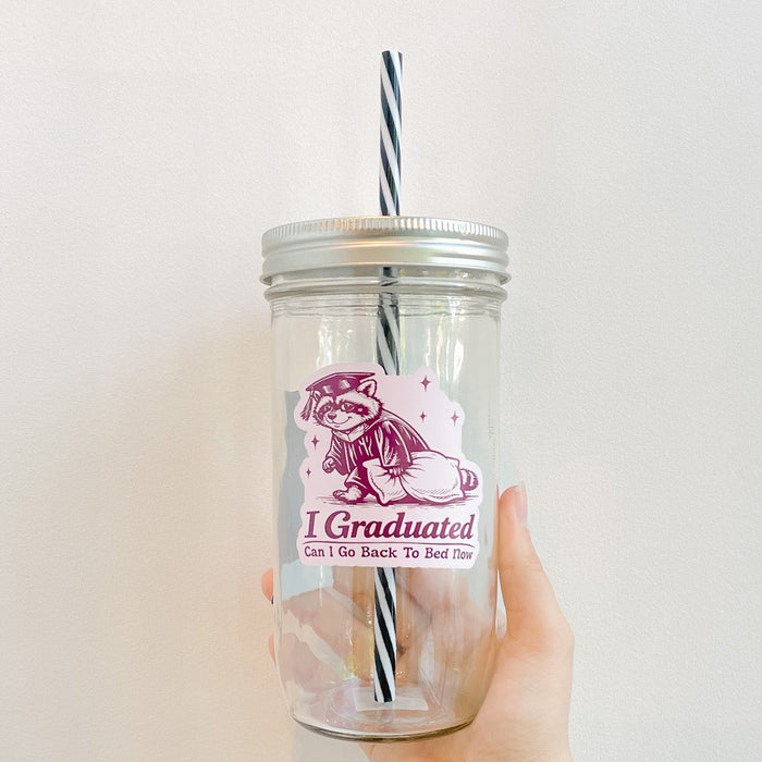 Hand holding a mason jar tumbler with multi-tone pink design of sleep raccoon with a graduation cap that reads i graduated, can i go back to bed now? With a black and white reusable striped straw and straw lid