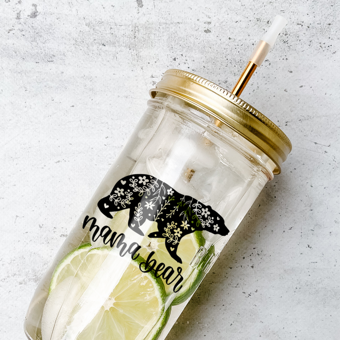 Mason jar tumbler filled with a citrus water drink and has a gold lid and straw and a calligraphy print that reads "mama bear" with white floral patterns in a black bear graphic. Photographed against a marble countertop.