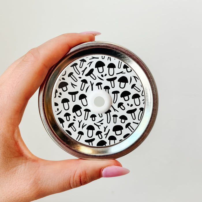 Hand holding mason jar patterned straw lid that has black and white shaped mushroom designs on white background