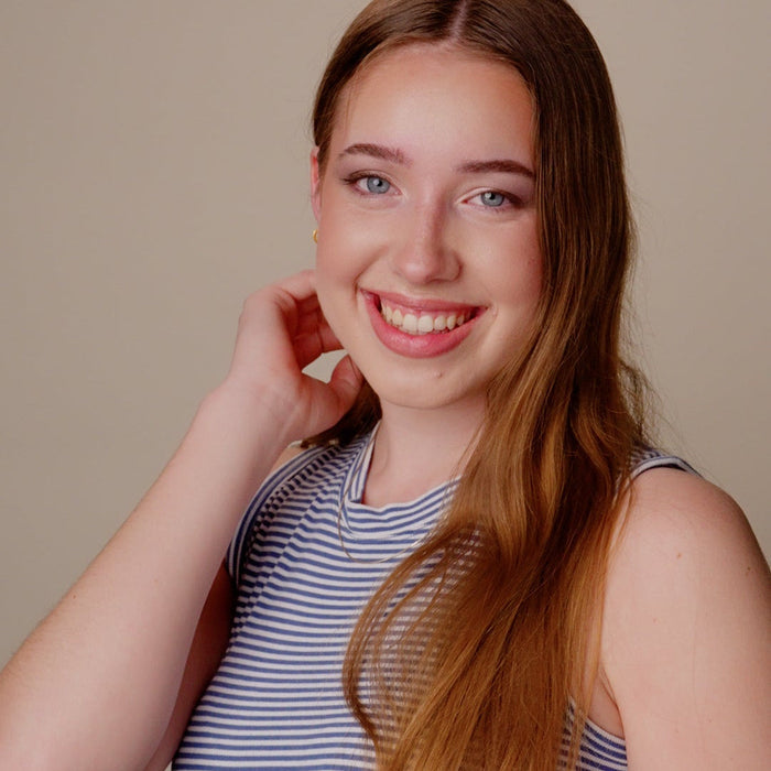 A smiling young woman with long light brown hair wearing a blue and white striped sleeveless top, posing against a solid beige background.