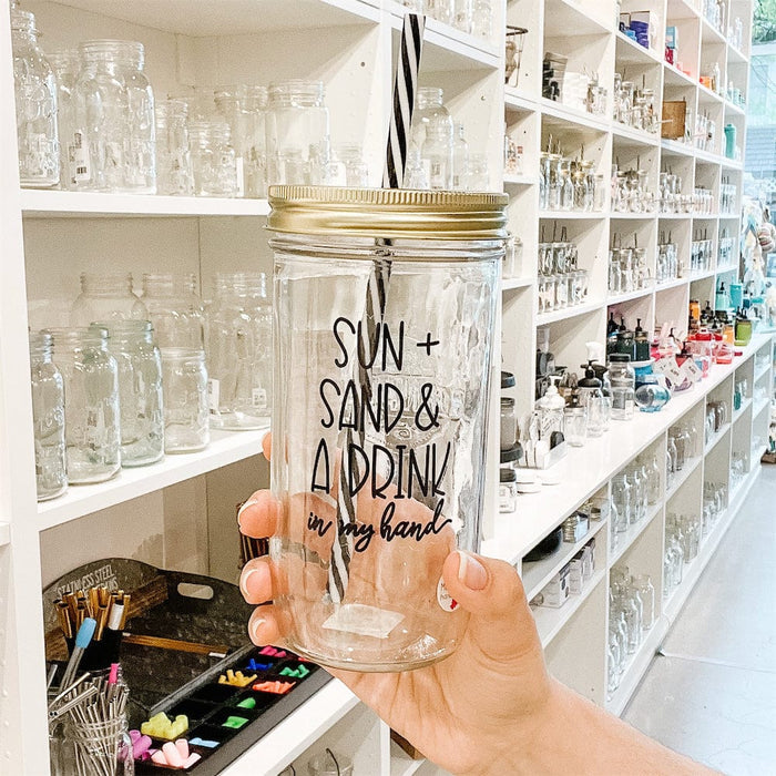 Photo of a mason jar tumblr with a sticker text that says "Sun + Sand & a Drink in my hand". It has gold lid with black and white straw.A photo of a mason jar tumbler with a sticker text that says "Sun + Sand & A Drink in my hand"