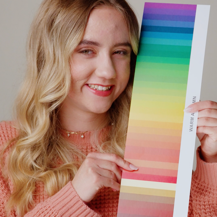 A smiling blonde woman in a peach knit sweater holds up a vertical color swatch card showing a rainbow gradient of colors against a plain light gray background.