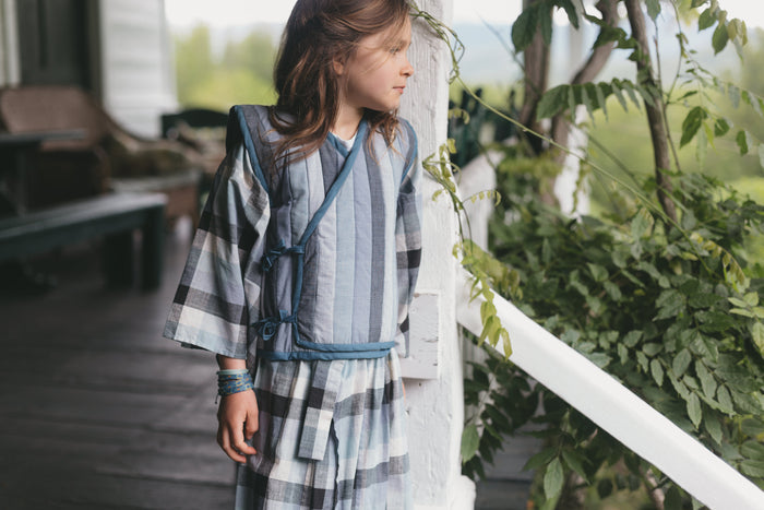 Young girl in a plaid kimono and vest standing on a porch with greenery in the background
