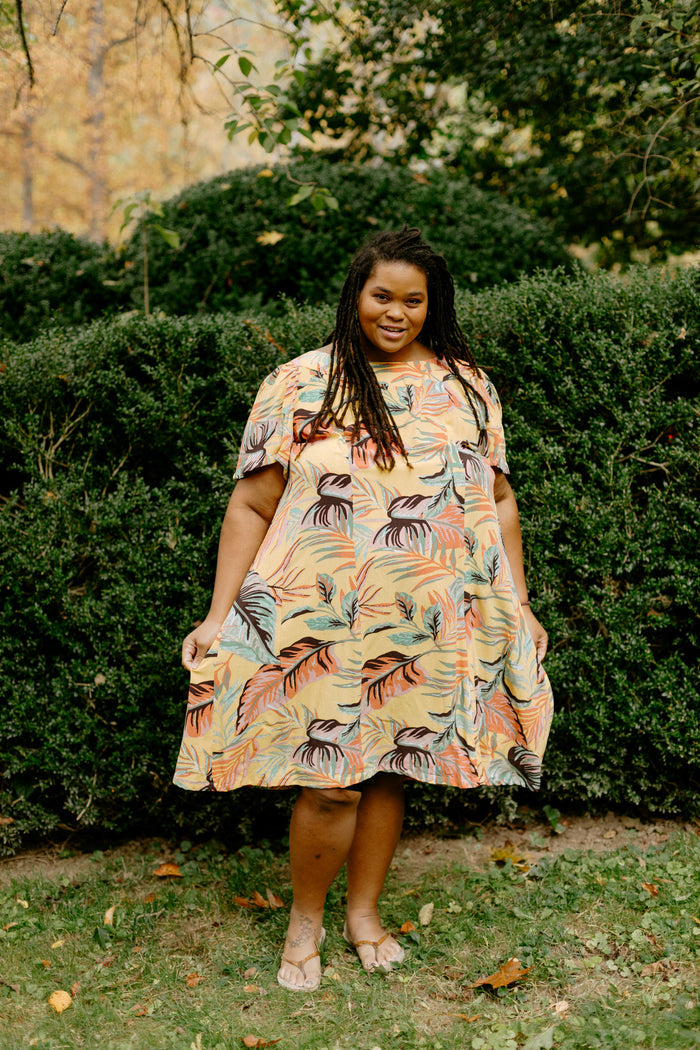 Black woman wearing a size 2XL knee-length, short-sleeve, yellow muumuu with orange and green palm fronds on it.