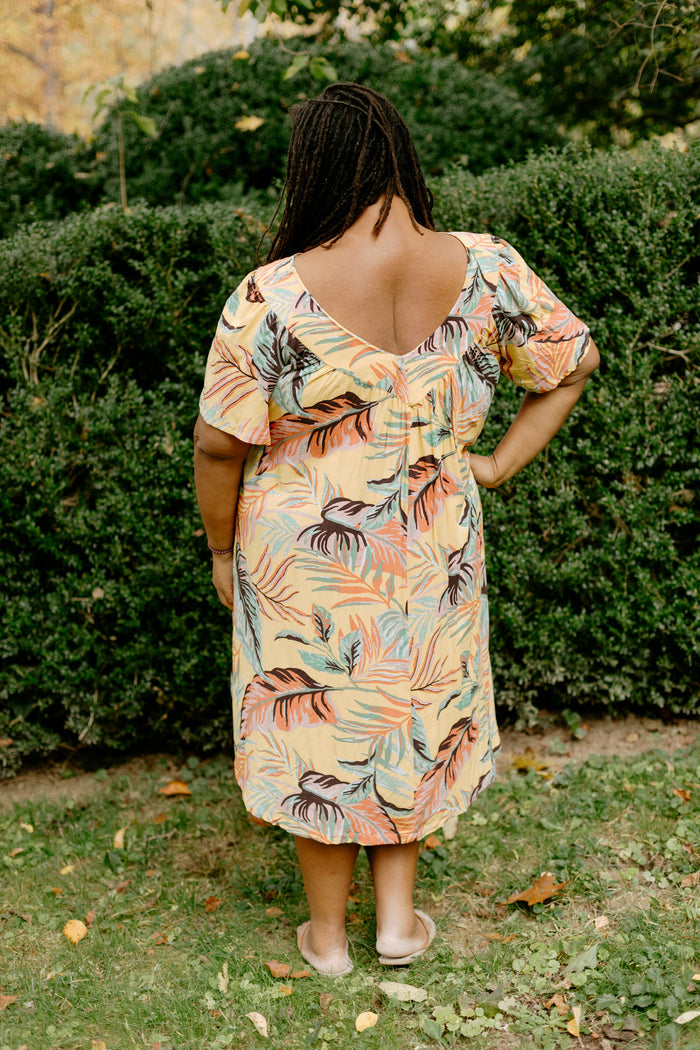 Back view of Black woman wearing a size 2XL knee-length, short-sleeve, yellow muumuu with orange and green palm fronds on it.