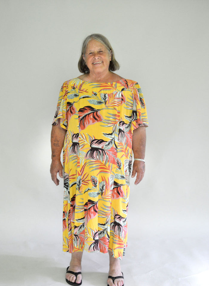 Older woman wearing a size 2XL knee-length, short-sleeve, yellow muumuu with orange and green palm fronds on it.