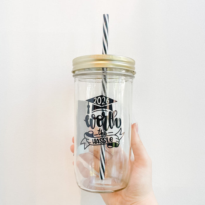 Hand holding a mason jar tumbler that reads in black text 2026 worth the hassle including a banner and grad cap design. Black and white reusable striped straw and straw lid.
