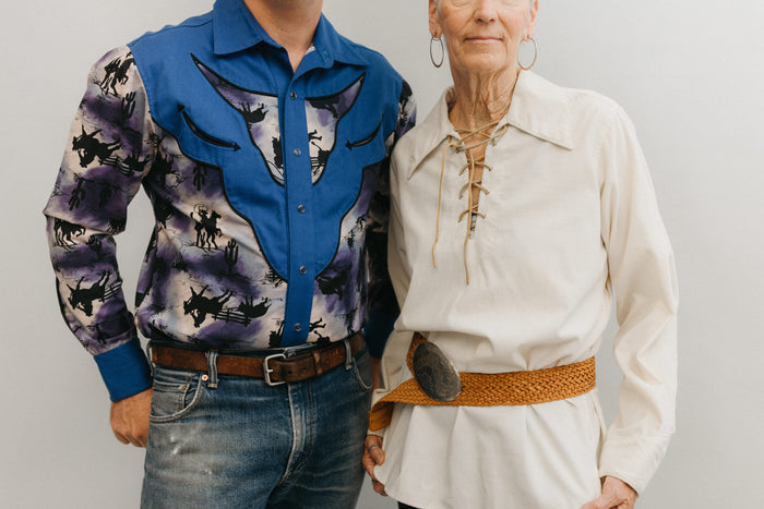 A man and a older woman standing in front of a white studio backdrop ,wearing 212 Five Frontier Shirts. Man wearing View D with decorative bull's head yoke and woman wearing View A with lace up front with belt around the waist.