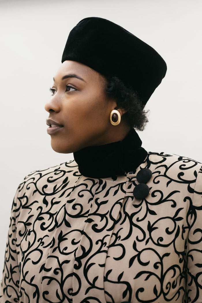 Head shot of a Black woman wearing a black velvet turban hat and a brown and black coat.