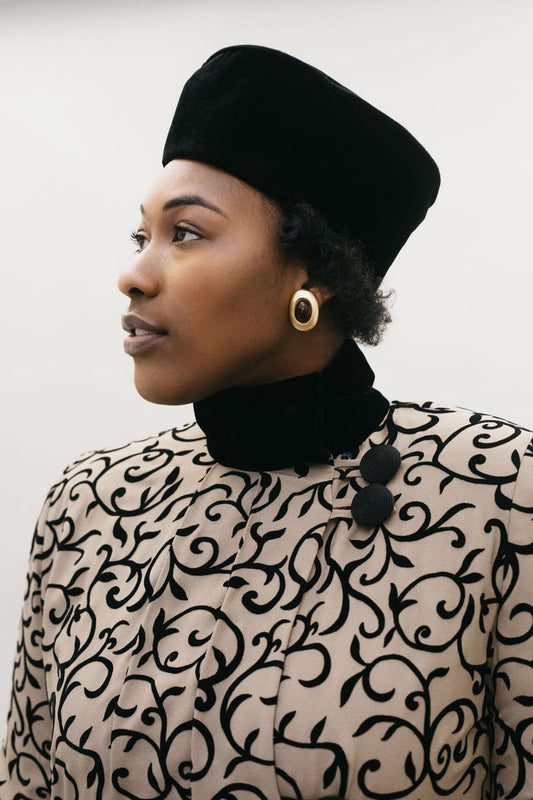 Head shot of a Black woman wearing a black velvet turban hat and a brown and black coat.