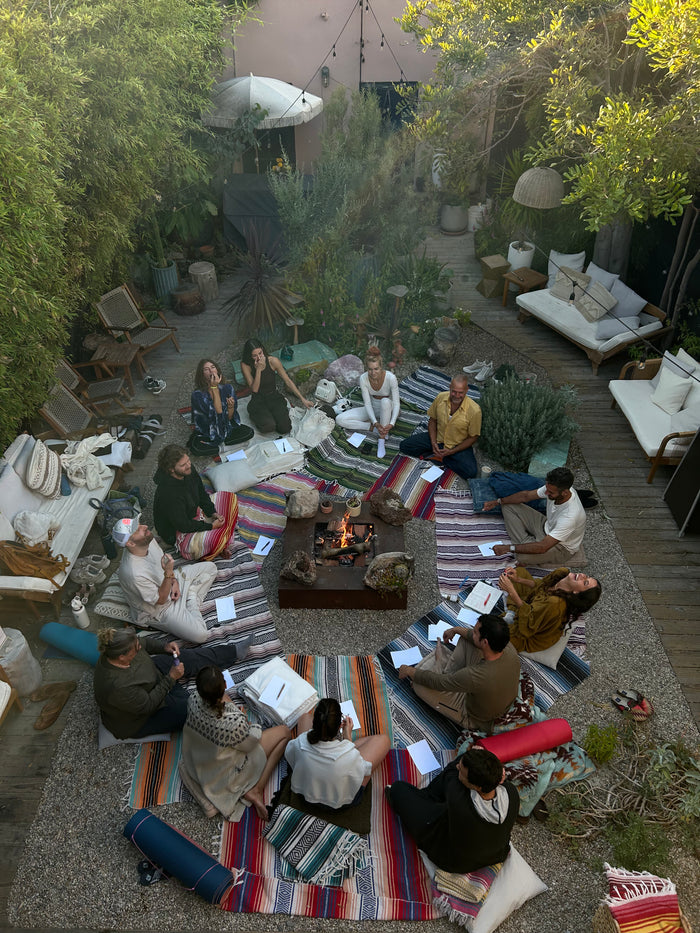 Group of people sitting on colorful blankets around a fire pit in a backyard setting.