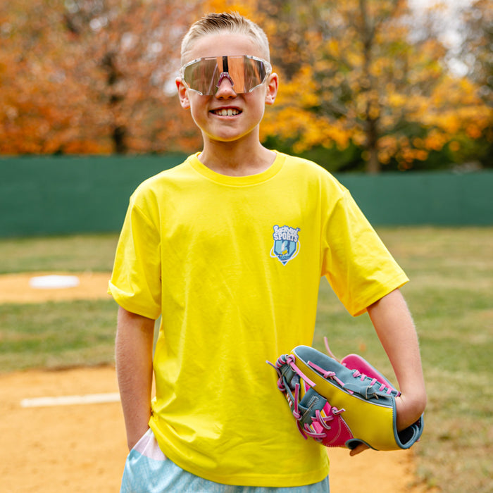 Young boy in a yellow 'BL101 Backyard Sports' t-shirt and sunglasses holding a baseball glove on a baseball field.