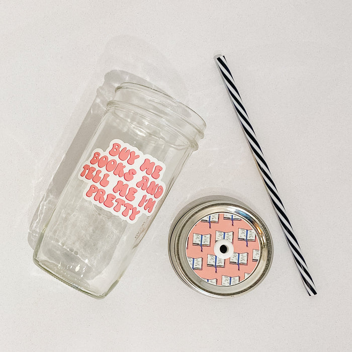 Flat lay of a mason jar tumbler that says “buy me books and tell me I’m pretty”, a pink straw lid with a repeating pattern of an opened book, and a black and white striped straw.