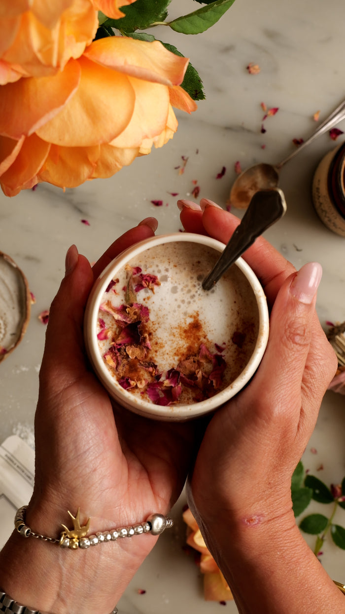 Two hands holding a small cup of coffee with a spoon, surrounded by flowers and a marble surface.