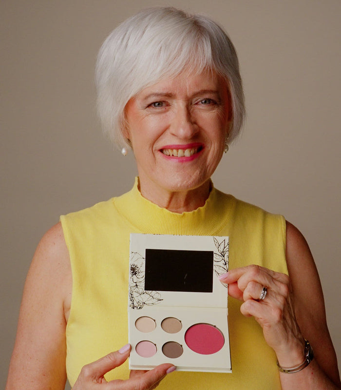 An older woman with short white hair, wearing a yellow sleeveless top, holds up a six-color eyeshadow palette with a mirror. The background is a solid neutral beige studio backdrop.