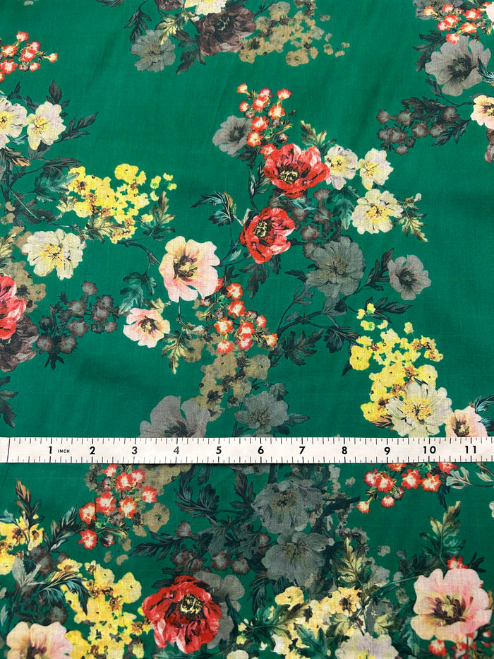 This is a cotton voile it has a vibrant and unsaturated floral print with yellow, pink and red flowers on a green background with a ruler for scale on a green background