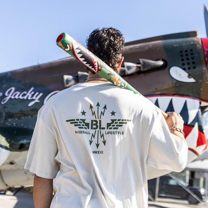 Person holding a baseball bat with a shark design, wearing a Tan t-shirt with dark green graphic featuring wings, arrows, and stars, with the Baseball Lifestyle BL logo in the center