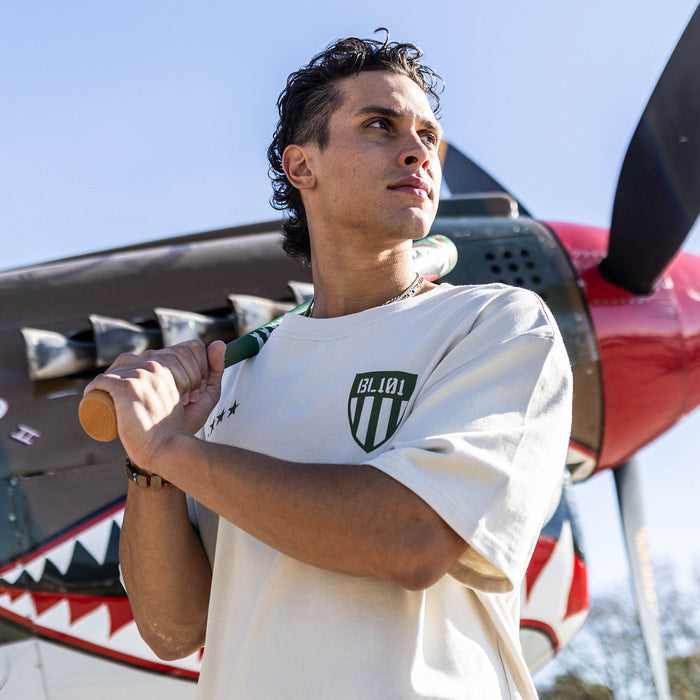 Person holding a baseball bat in front of a red airplane wearing a Tan t-shirt with dark green graphic with 4 stars and armor BL101 logo