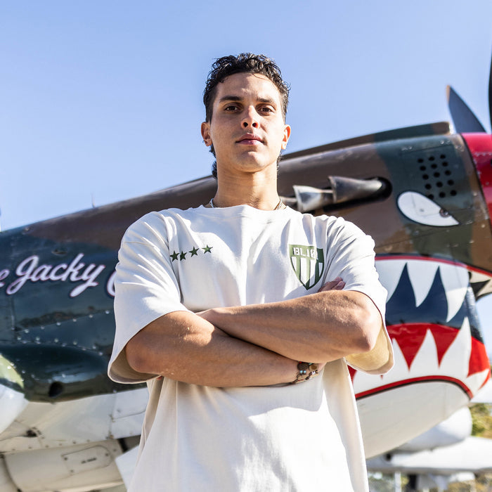 Person wearing Tan t-shirt with dark green graphic with 4 stars and armor BL101 logo in front of a airplane.