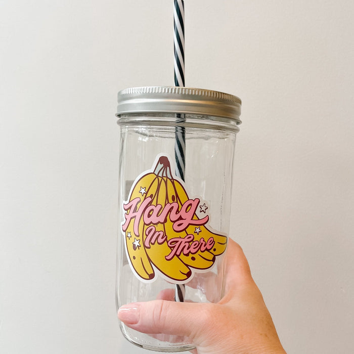 Hand holding a mason jar tumbler that says “hang in there” in pink lettering over a bunch of bananas.