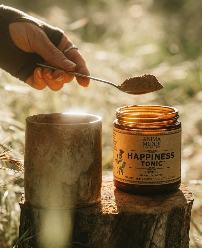 A hand holds a spoonful of brown powder above an amber glass jar labeled Anima Mundi Herbs Happiness Tonic, set on a rustic wooden stump outdoors with warm sunlight and a blurred natural background.
