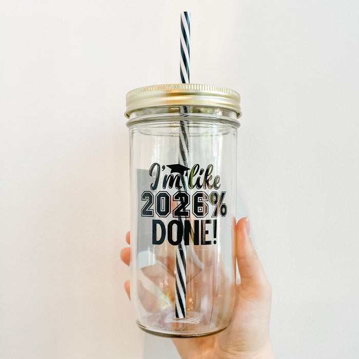 Hand holding a glass mason jar tumbler that reads in black text I’m like 2026% done! With a little grad cap design. Black and white striped reusable straw and straw lid.