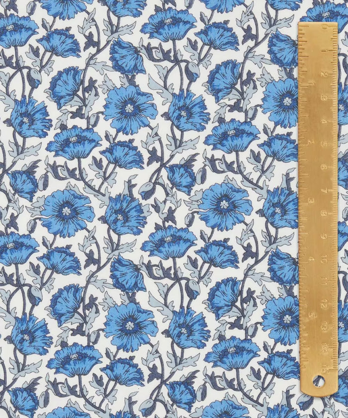 Liberty of London cotton tana lawn in a  beautiful, vibrant print of blue poppies with a white background. With a light brown ruler on top.