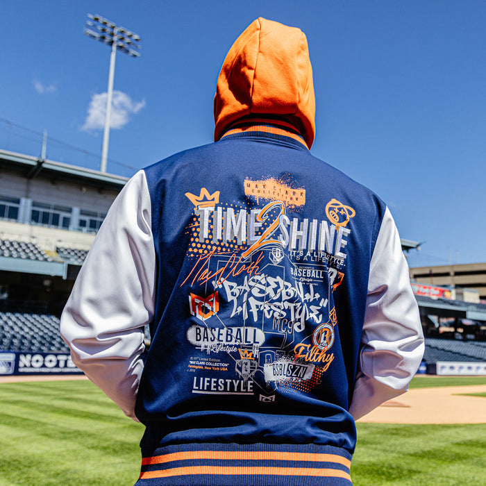 Person wearing a blue letterman jacket with orange and white accents on a baseball field.