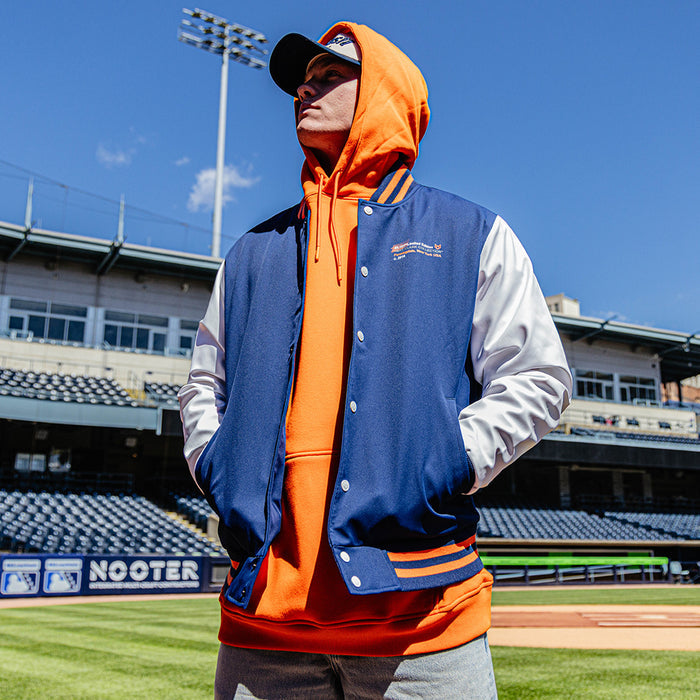 Person wearing a blue and orange letterman jacket with a hood on a baseball field.
