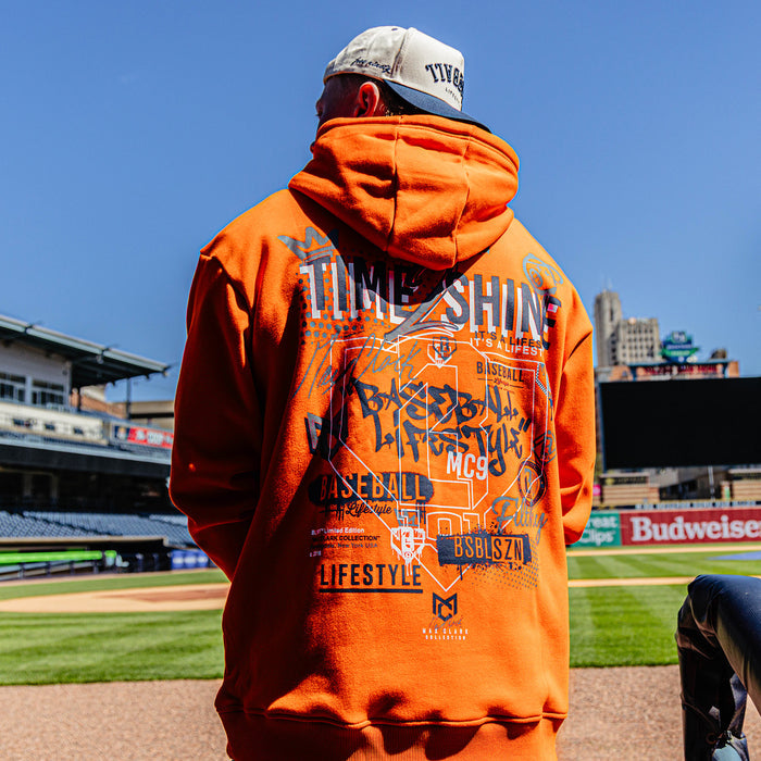 Person wearing an orange hoodie with graphics at a baseball stadium
