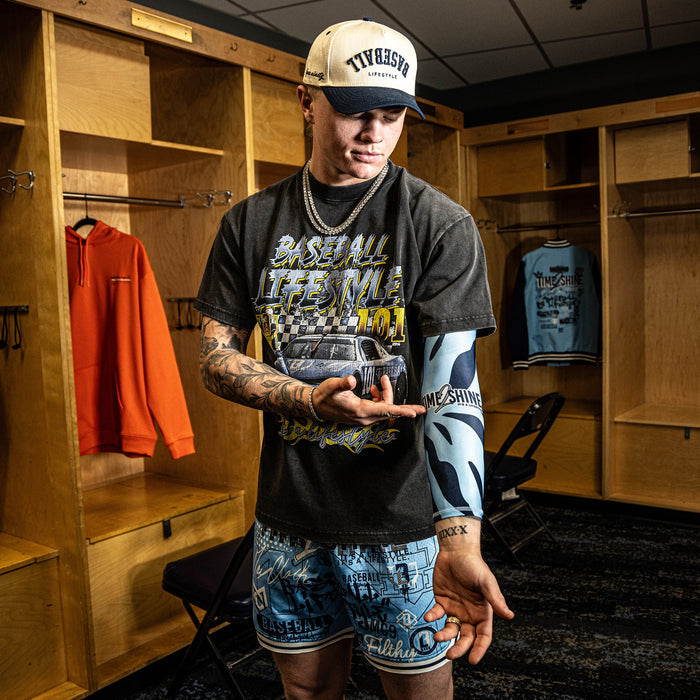 Man in a locker room wearing a graphic t-shirt and shorts, holding a baseball.