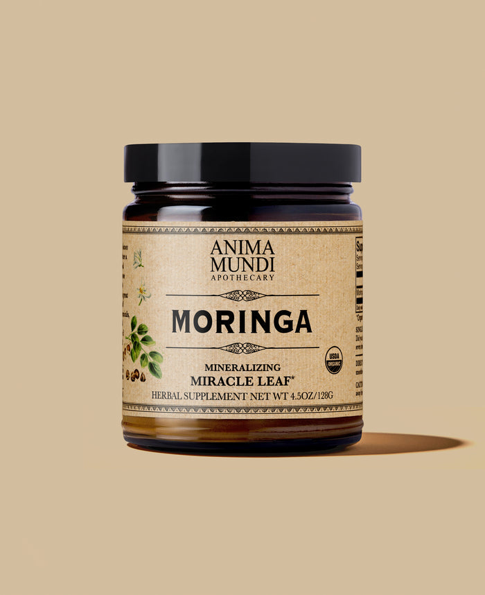 A jar of Anima Mundi Apothecary Moringa Mineralizing Super-Tonic herbal supplement, featuring a black lid and a textured beige label with botanical illustrations, set against a solid beige background.
