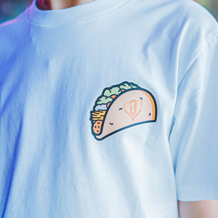 Person wearing a white t-shirt with a taco graphic in front of a colorful neon sign.
