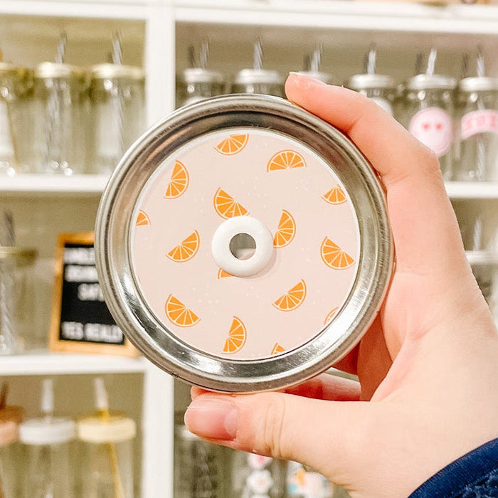 Hand holding a mason jar patterned straw lid with little orange slices on a light peach background