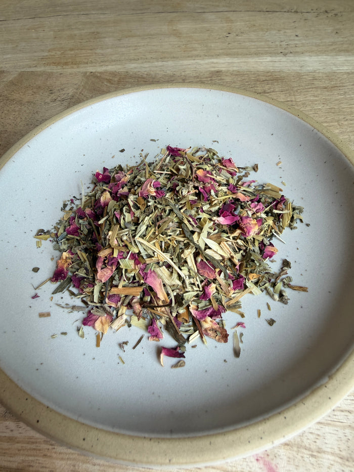 A pile of loose-leaf herbal tea blend with dried rose petals and green leaves, presented in a small beige ceramic bowl on a light wood surface.