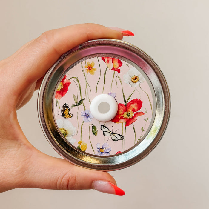 straw lid with a pink background, there are green stems/leaves taking up most of the centre of the lid, there are also flowers and butterflies scattered