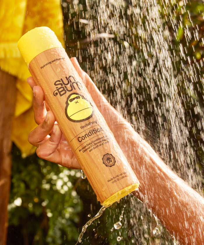 A hand holding a yellow-capped bottle of Sun Bum conditioner under a stream of water outdoors with green foliage in the background.