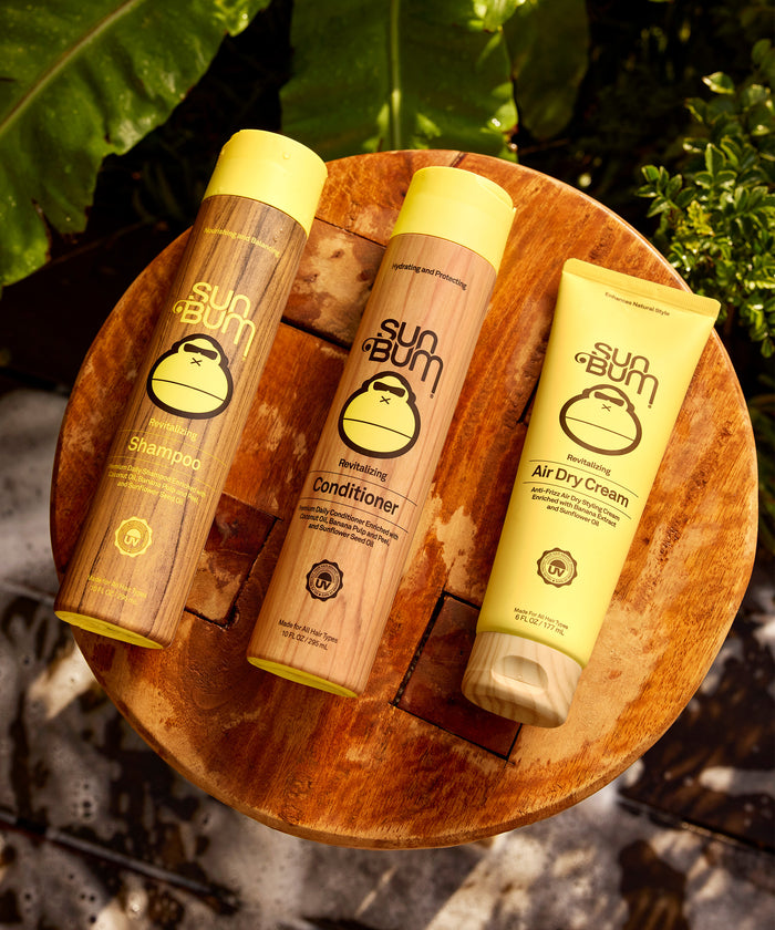 Three Sun Bum hair care products, including shampoo, conditioner, and air dry cream, arranged on a round wooden surface outdoors with green foliage in the background.