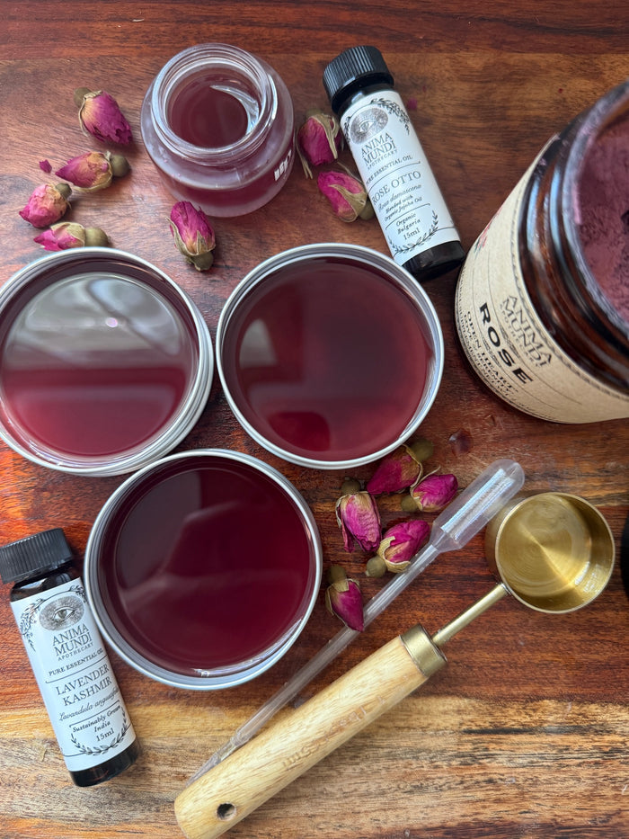 Spice jars, a bottle of lavender candy, and a wooden spoon on a wooden surface with rose petals.