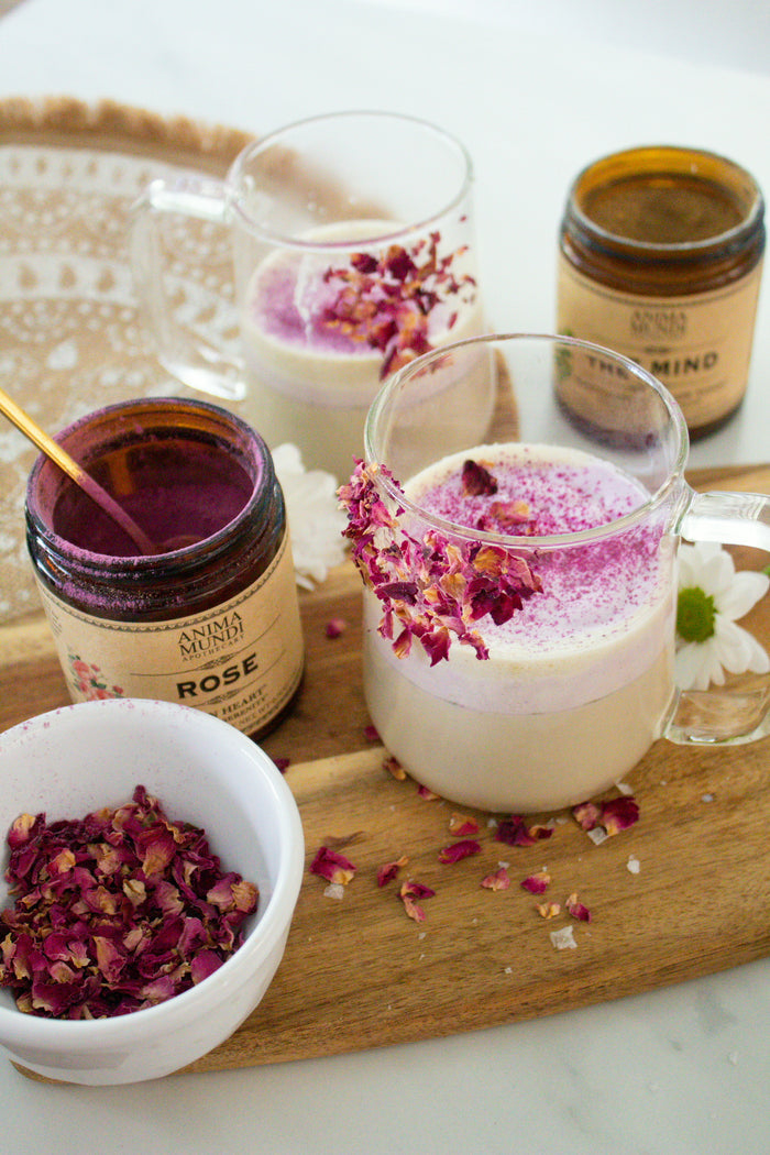 Mugs with pink and white beverages on a wooden tray with rose petal jars and a bowl of rose petals.
