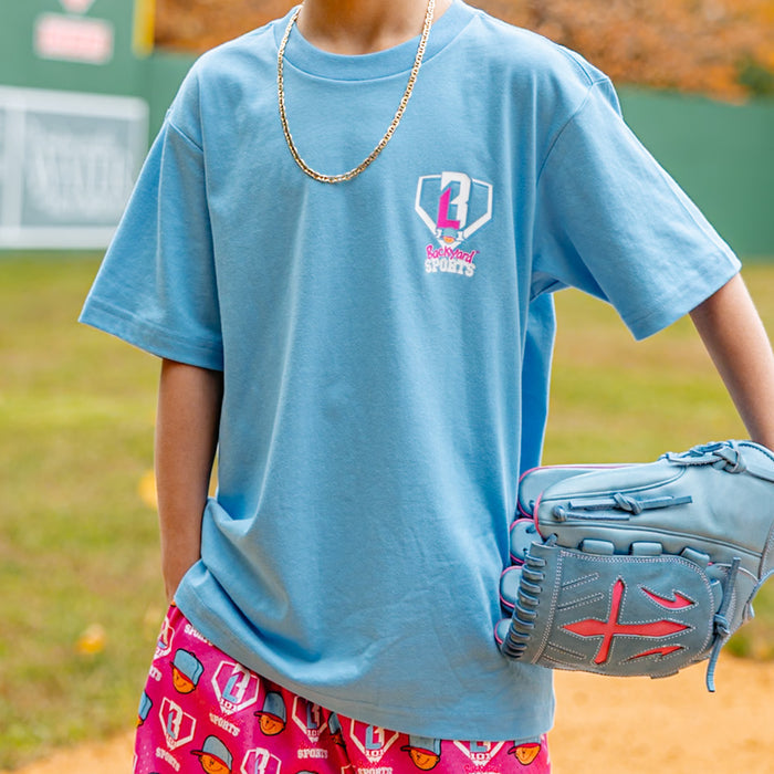 Person wearing a blue t-shirt with a 'BL101 Backyard Sports' logo and pink 'Pablo Sanchez' shorts, holding a baseball glove on a field.