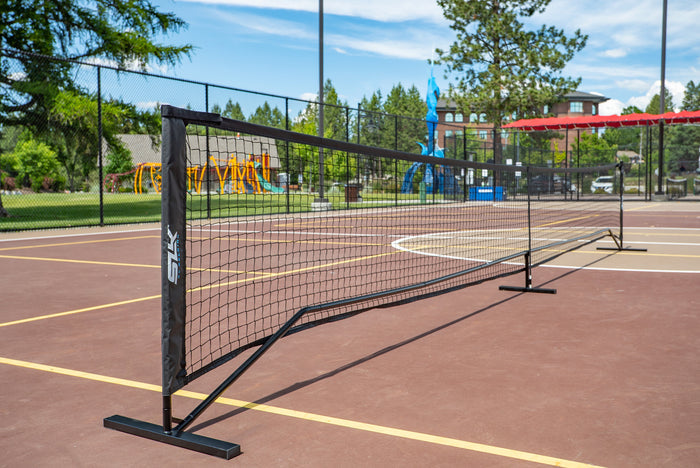 Portable pickleball net on an outdoor multiuse court with yellow lines; playground and trees in background. SLK on post