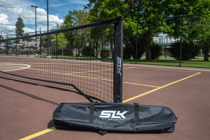 Portable SLK-branded net bag on outdoor hardcourt beside a raised net, with trees and buildings in background.