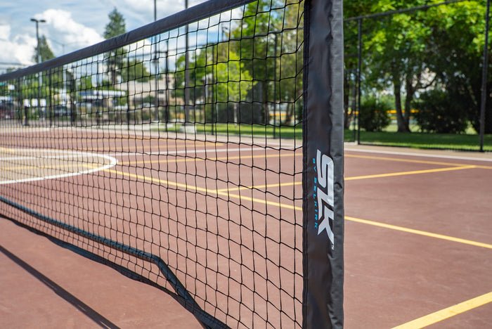 Tennis net across an outdoor brown court with yellow lines; black net post sleeve labeled 'SLK' in foreground.