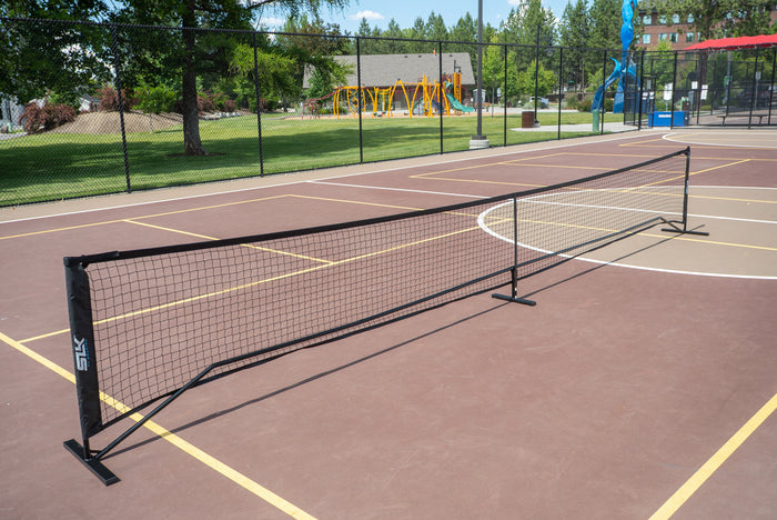 Portable SKLZ low-profile pickleball/tennis net set up on an outdoor multipurpose court with playground in background.