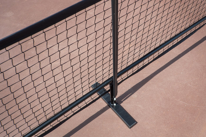 Portable metal mesh barrier with vertical post and T-shaped foot mounted on a reddish court surface.