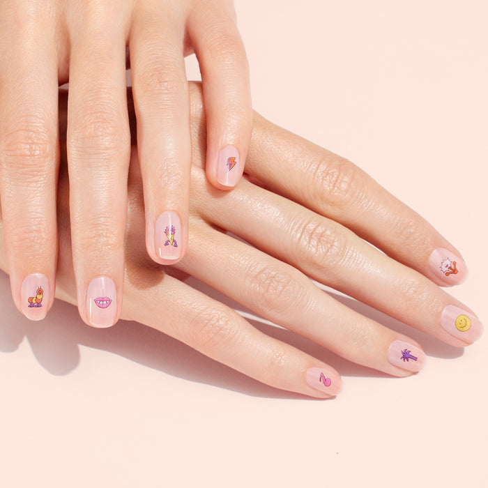 Close-up of hands with short, natural nails, each decorated with a different playful Summer Nail Art Sticker from Manucurist—smiley faces, lips, and more—over a light pink base for a fun manicure.